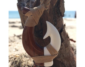 May include: A handcrafted pendant necklace featuring a fish hook design. The hook is made of dark brown wood and white material, wrapped with tan cord. The necklace hangs from a thick, braided rope. The text "MAORI MAN" is visible at the bottom.