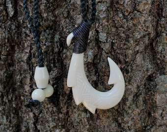 May include: A white bone fish hook pendant on a black cord necklace. The hook is carved with a traditional design and has a smooth, polished finish.