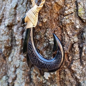May include: A handcrafted black pendant necklace featuring a fish hook design. The hook is carved with intricate patterns and is suspended from a woven beige cord. The background is a textured tree trunk.