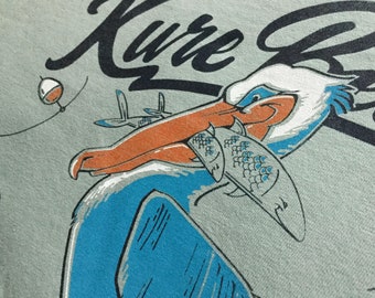 Puede incluir: Un pelícano azul y blanco con un pico naranja sostiene un pez en su pico. El pelícano está sobre un fondo gris claro con el texto "Kure Beach" en negro.