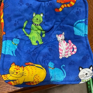 May include: A blue fabric bowl cozy with a quilted design. The cozy is decorated with colorful cartoon cats in various poses. The cats are yellow, green, pink, and blue, with white accents. The background has paw prints.