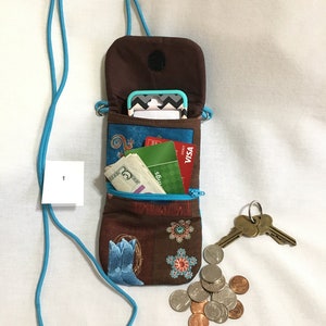 May include: A brown and blue fabric crossbody phone pouch with a zipper closure. The pouch is open and contains a mobile phone, cash, credit cards, and keys. The pouch has a turquoise strap.