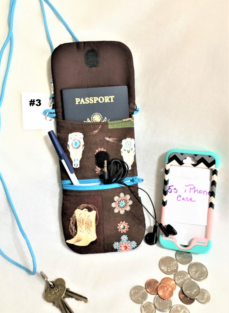 May include: A brown fabric phone case with a zipped pocket and a skull and cowboy boot design. The case is open and a passport is visible inside. A blue pen, a pair of headphones, and a key chain with keys are also visible. The case is hanging from a blue lanyard.