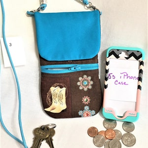May include: A turquoise crossbody phone purse with a brown fabric lining featuring a cowboy boot design. The purse has two zippered compartments. A turquoise phone case with a black and white chevron pattern is next to the purse. The text "5s iPhone Case" is written on the phone case. There are also keys and coins on the white surface.