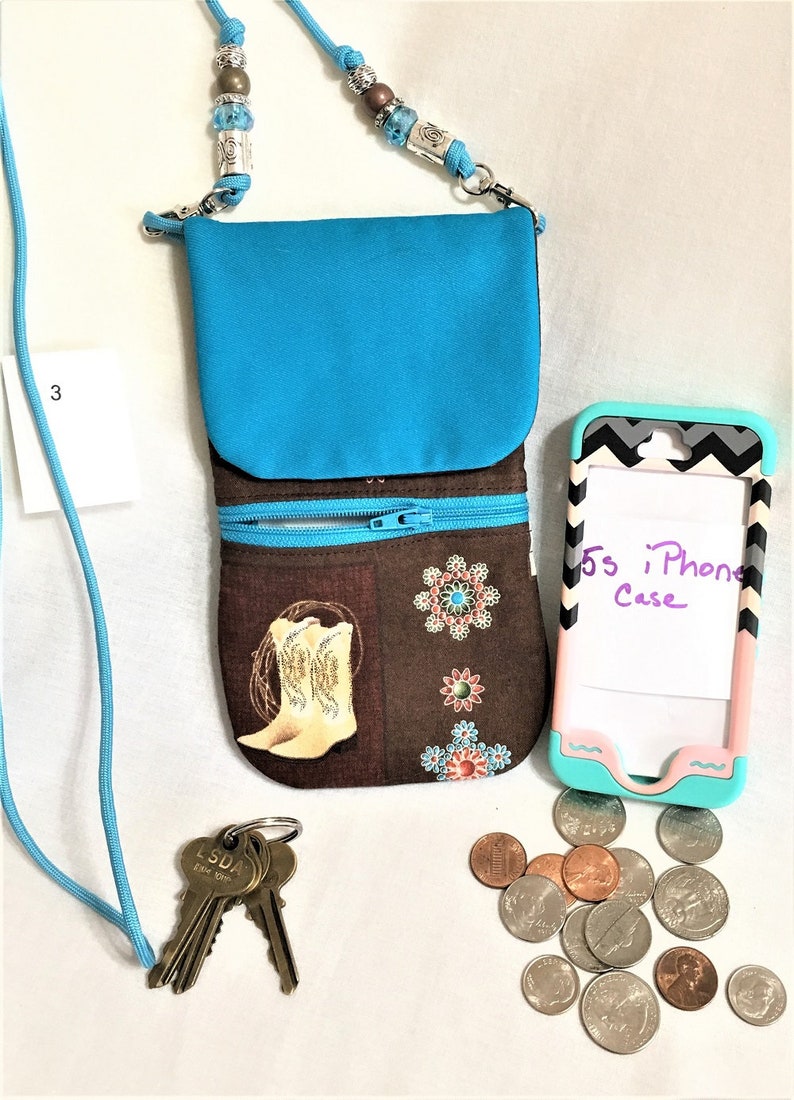 May include: A turquoise crossbody phone purse with a brown fabric lining featuring a cowboy boot design. The purse has two zippered compartments. A turquoise phone case with a black and white chevron pattern is next to the purse. The text "5s iPhone Case" is written on the phone case. There are also keys and coins on the white surface.