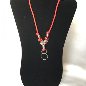 May include: A red lanyard with a silver ring and red and silver beads. The lanyard is displayed on a black mannequin.