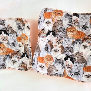May include: Two quilted pot holders featuring a colorful cat print. The larger pot holder is orange, white, and gray with a variety of cat faces. The smaller pot holder is the same print but in a smaller size.
