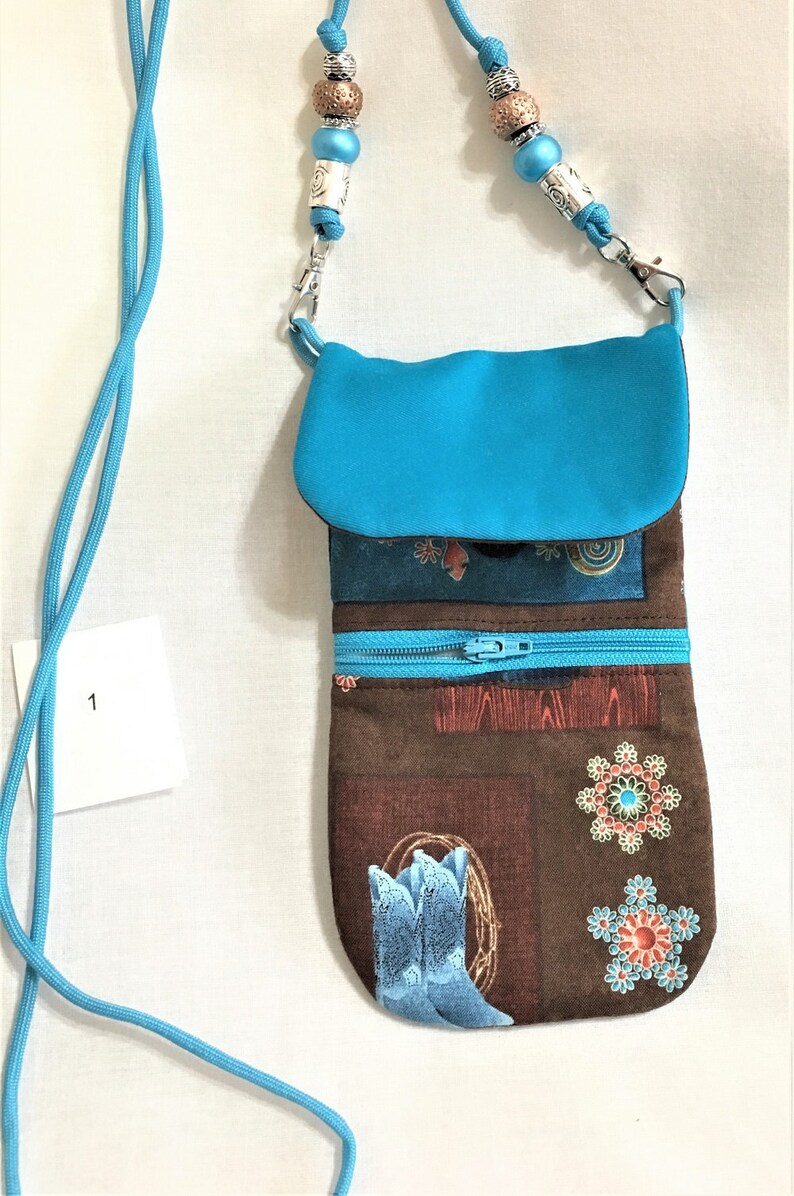 May include: A turquoise blue fabric pouch with a zipper closure and a brown fabric base with a western-themed print featuring boots, flowers, and geometric patterns. The pouch has a long turquoise blue strap with silver beads.