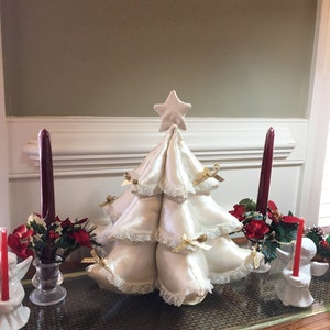 Könnte beinhalten: Ein cremefarbener Weihnachtsbaum mit Sternspitze, verziert mit goldenen Schleifen und Spitzensaum. Rote Kerzen und Blumengestecke flankieren den Baum und schaffen eine festliche Weihnachtsdekoration.