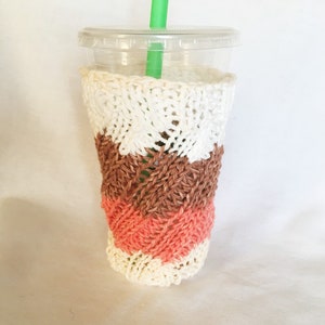 May include: A white, brown, and pink knitted cup cozy with a diagonal pattern. The cozy is on a clear plastic cup with a green straw.