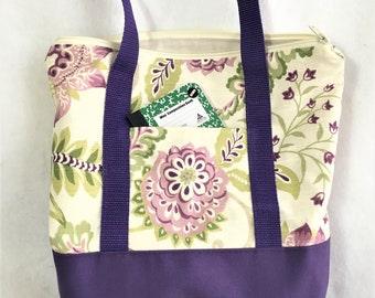 May include: A floral print tote bag with a purple base and straps. The bag features a cream-colored background with a pattern of pink and green flowers. A small notebook is visible in the front pocket. The bag has a zipper closure.