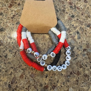 Peut inclure: Ensemble de quatre bracelets en perles avec les mots "Ohio", "Go", "Buckeyes" et "Yes" écrits en lettres noires sur des perles blanches. Les bracelets sont faits de perles rouges, blanches et grises.