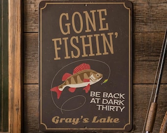 以下が含まれることがあります： 魚のイラストが描かれた「Gone Fishin'」の看板がある素朴な風景。 釣り竿、タックルボックス、ランタン、柳のバスケットが木製の表面に配置されています。 看板には「Be Back at Dark Thirty」と「Gray's Lake」と書かれています。