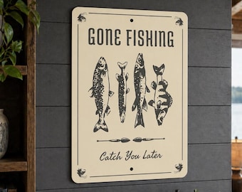 以下が含まれることがあります： 「GONE FISHING」の文字と「Catch You Later」の文字が上下に配置された長方形の金属製の看板。クリーム色の背景に、4匹の様式化された魚のイラストが黒色で描かれています。看板は濃い灰色の壁に取り付けられています。