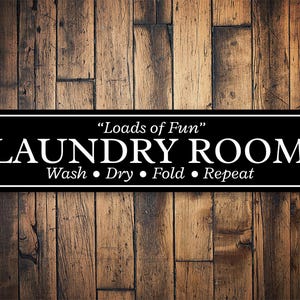 May include: Black and white sign with the text "Loads of Fun" Laundry Room Wash, Dry, Fold, Repeat on a wood background.