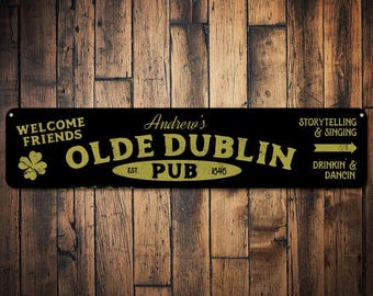 Puede incluir: Un letrero negro y dorado con el texto "Welcome Friends Andrew's Olde Dublin Pub Est. 1846" y flechas apuntando a la derecha con el texto "Storytelling & Singing" y "Drinkin & Dancin".