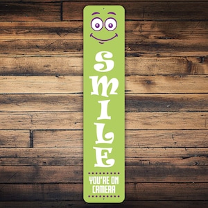 May include: A green metal sign with a white smiling face and the word "SMILE" in white letters. The sign also has the text "YOU'RE ON CAMERA" in purple letters.