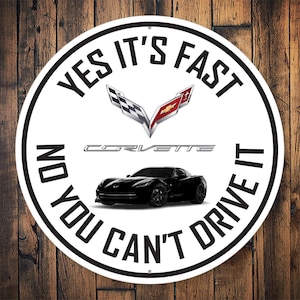 May include: A white round metal sign with black text that reads "Yes it's fast no you can't drive it". The sign also features a black and white chequered flag and a red Corvette logo. A black Corvette is pictured in the centre of the sign.