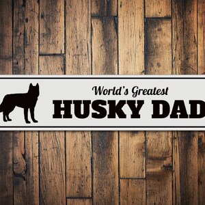 May include: White metal sign with black text and a silhouette of a husky dog. The sign reads "World's Greatest Husky Dad."