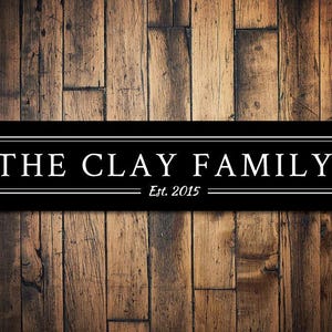 以下が含まれることがあります： 木製の背景に「The Clay Family Est. 2015」という文字が書かれた白黒の看板です。