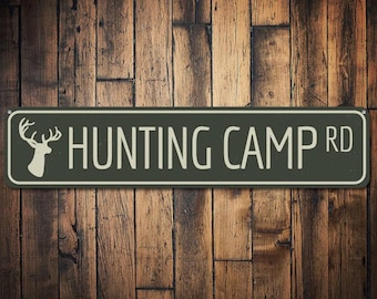 May include: A dark green metal sign with white text and a white silhouette of a deer head. The sign reads "Hunting Camp Rd."