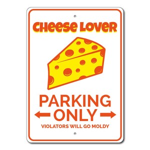 May include: A white metal sign with orange text that reads "Cheese Lover Parking Only" with an arrow pointing to the left and an arrow pointing to the right. Below the text is a smaller orange text that reads "Violators Will Go Moldy". A cartoon image of a yellow wedge of cheese with orange spots is above the text.