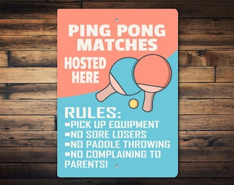 May include: A rectangular sign with a wooden background, featuring the text "PING PONG MATCHES HOSTED HERE." The sign has a peach and blue color scheme with illustrations of ping pong paddles and a ball. The rules are listed below.