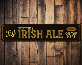 Puede incluir: Un letrero negro y dorado con el texto "Try Scottie's Irish Ale On Tap Here" y "Brewed in Dublin" en una fuente más pequeña.