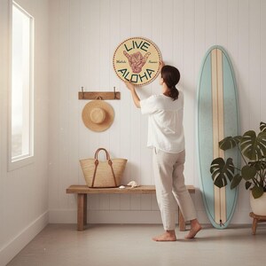 A woman hangs a round sign with the word "ALOHA" on a wall in a room with a wooden bench, a straw hat, and a surfboard.