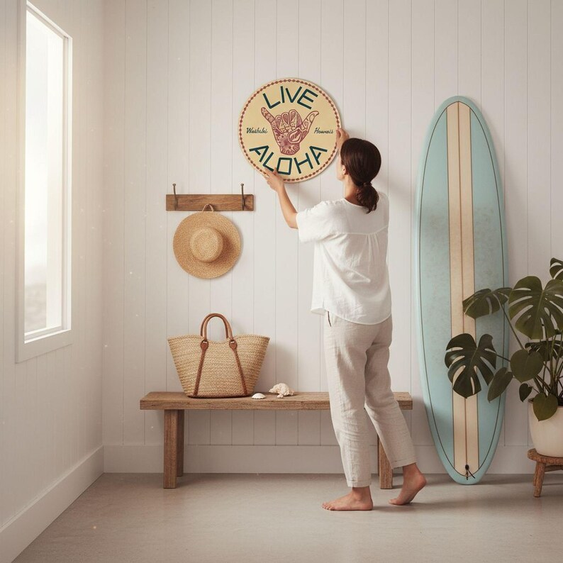 A woman hangs a round sign with the word "ALOHA" on a wall in a room with a wooden bench, a straw hat, and a surfboard.