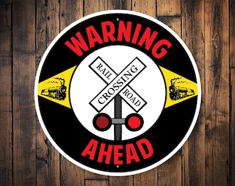 Può includere: Un cartello rotondo nero e giallo con la scritta "WARNING" in lettere rosse e "AHEAD" in lettere rosse. Il cartello presenta anche un cartello di attraversamento ferroviario bianco con la scritta "RAIL CROSSING ROAD" e un'illustrazione in bianco e nero di un treno con fumo che esce dal camino.