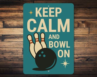 May include: A teal metal sign with the words "KEEP CALM AND BOWL ON" in white. The sign features a bowling ball and pins in a retro style. A starburst graphic is in the lower right corner.