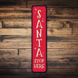 May include: A red metal sign with white lettering that reads "SANTA STOP HERE" with snowflakes around the letters.