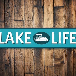 May include: A teal blue metal sign with white text that reads "LAKE LIFE". A white silhouette of a pontoon boat is in a circle in the middle of the sign.