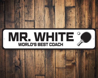 Könnte beinhalten: Weißes Metallschild mit schwarzem Text "MR. WHITE WORLD'S BEST COACH" und einem schwarz-weißen Tischtennis Schläger mit einem weißen Ball.
