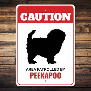 May include: White metal sign with a red border and a black silhouette of a dog. The sign reads "CAUTION" and "AREA PATROLLED BY PEEKAPOO".