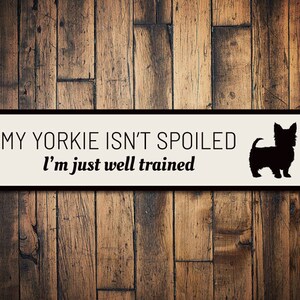 May include: A white sign with black text and a black silhouette of a Yorkshire Terrier. The sign reads "My Yorkie isn't spoiled I'm just well trained."