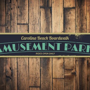 Puede incluir: Un letrero de estilo vintage con borde dorado y fondo negro. El letrero dice "Carolina Beach Boardwalk Amusement Park" en texto blanco con contorno verde. El texto "Rides Open Daily" está en texto blanco más pequeño debajo del texto principal.