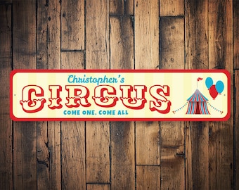 May include: A red and white circus sign with the text "Christopher's Circus Come One, Come All" in a vintage font. The sign also features a red and blue striped tent with a red flag and blue balloons.