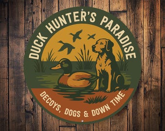 Op de afbeelding: Rond bord met een jachtthema. Het bord toont een eend in het water, een hond en vliegende eenden tegen een zonsondergang. De tekst luidt "Duck Hunter's Paradise, Decoys, Dogs & Down Time."