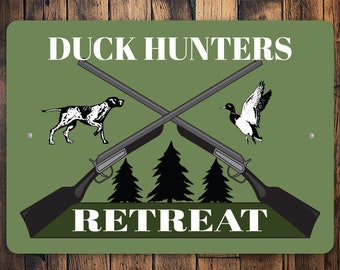 Może przedstawiać: Zielony metalowy znak z białym napisem "Duck Hunters Retreat". Znak przedstawia parę skrzyżowanych strzelb, lecącego kaczora i psa myśliwskiego.