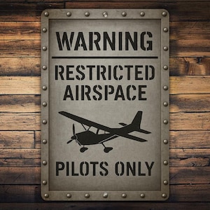 May include: A rectangular metal sign with the words "WARNING RESTRICTED AIRSPACE PILOTS ONLY" in black stencil font. A black silhouette of a small airplane is below the text. The sign is mounted on a wooden background.