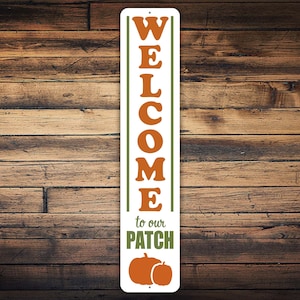 Puede incluir: Un letrero de metal blanco con texto verde y naranja que dice "WELCOME to our PATCH". El letrero presenta dos calabazas naranjas.