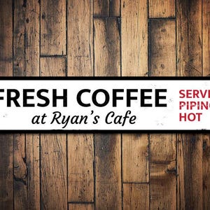 May include: White metal sign with black text that reads "FRESH COFFEE at Ryan's Cafe" and red text that reads "SERVED PIPING HOT".