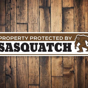 May include: White sign with brown lettering that reads "Property Protected by Sasquatch". A cartoon Sasquatch is illustrated on the right side of the sign.