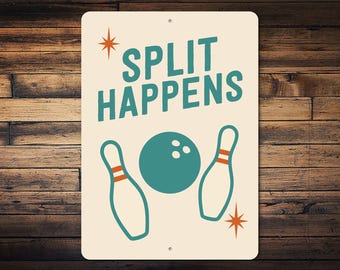 May include: A rectangular sign featuring the text "SPLIT HAPPENS" in teal, a teal bowling ball, and two bowling pins. The sign has a cream background and orange starburst accents. It is set against a wooden background.