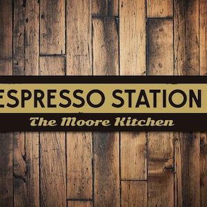May include: A rectangular sign with the words "ESPRESSO STATION" in gold letters and an arrow pointing right. Below, in smaller gold text, it reads "The Moore Kitchen." The sign is dark brown and mounted on a wooden background.