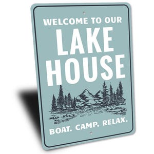 May include: A rectangular sign with a light blue background and white text that reads "WELCOME TO OUR LAKE HOUSE." Below the text is a black and white illustration of mountains, trees, and water. The bottom of the sign says "BOAT. CAMP. RELAX."