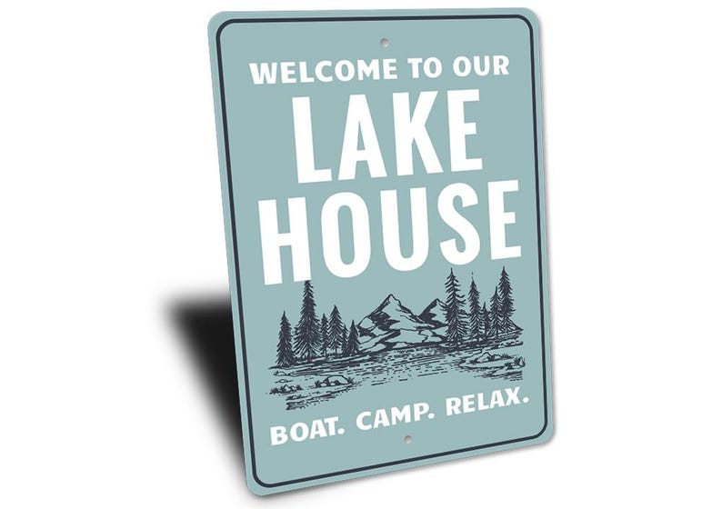 May include: A rectangular sign with a light blue background and white text that reads "WELCOME TO OUR LAKE HOUSE." Below the text is a black and white illustration of mountains, trees, and water. The bottom of the sign says "BOAT. CAMP. RELAX."