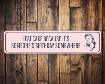 May include: Pink metal sign with the text "I eat cake because it's someone's birthday somewhere" and a retro illustration of a woman with a smile and a finger pointing to the right.